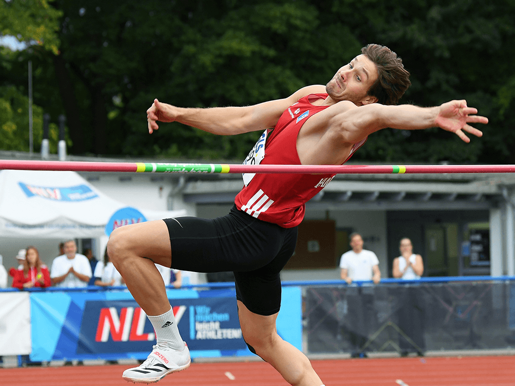 An athlete in a red uniform attempts a high jump, bending back to clear the bar, with spectators and greenery in the background.