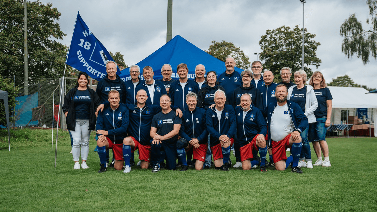 A group of people in sports attire pose together outdoors, with a blue tent and flag in the background, standing on green grass.