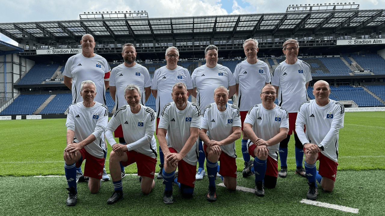 A football team of twelve men in white jerseys and red shorts posing on a stadium field with empty blue stands in the background.