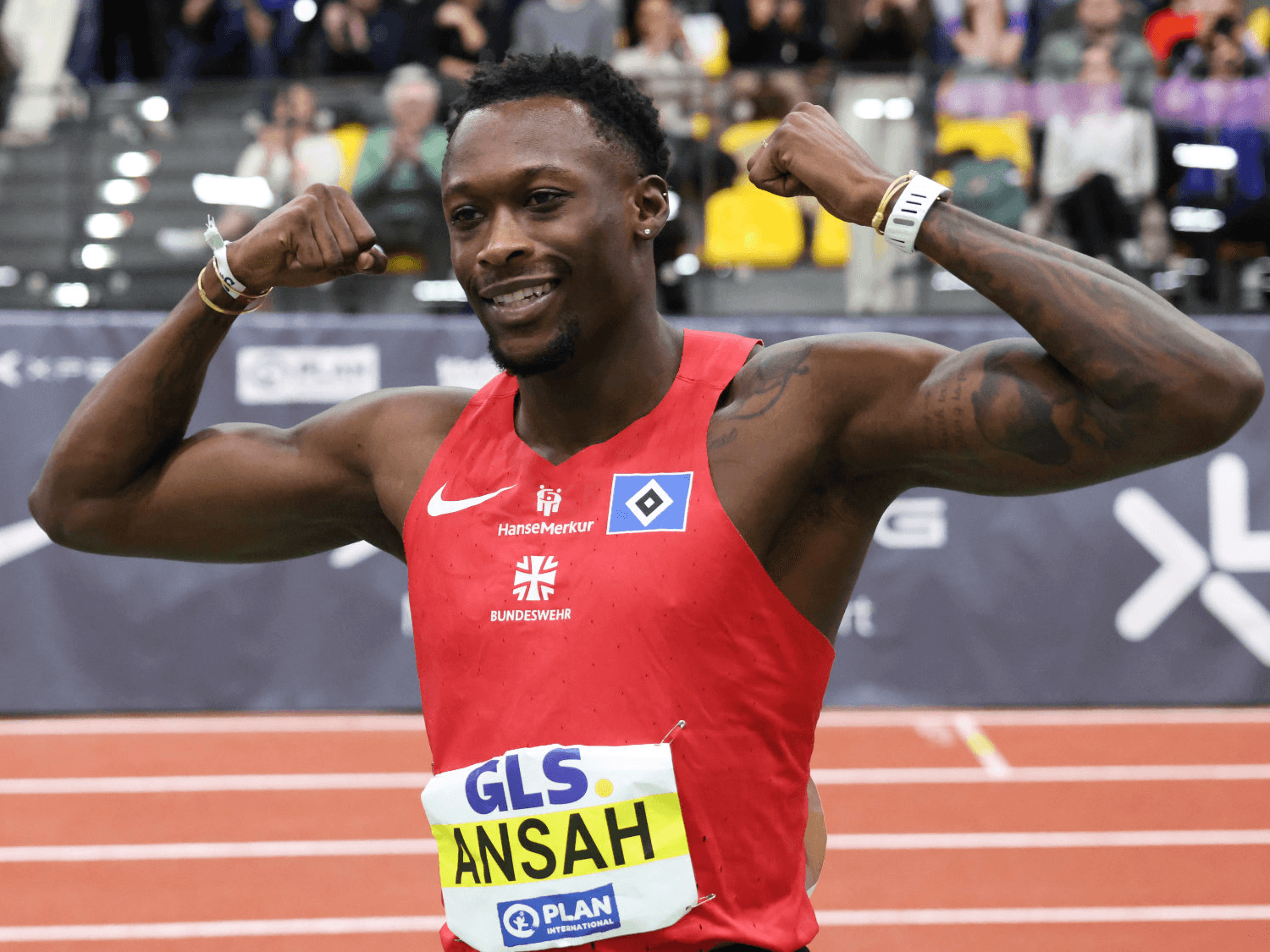 Athlete in a red jersey celebrates by flexing muscles on a track, wearing a race bib labeled "ANSAH," with a cheering crowd in the background.