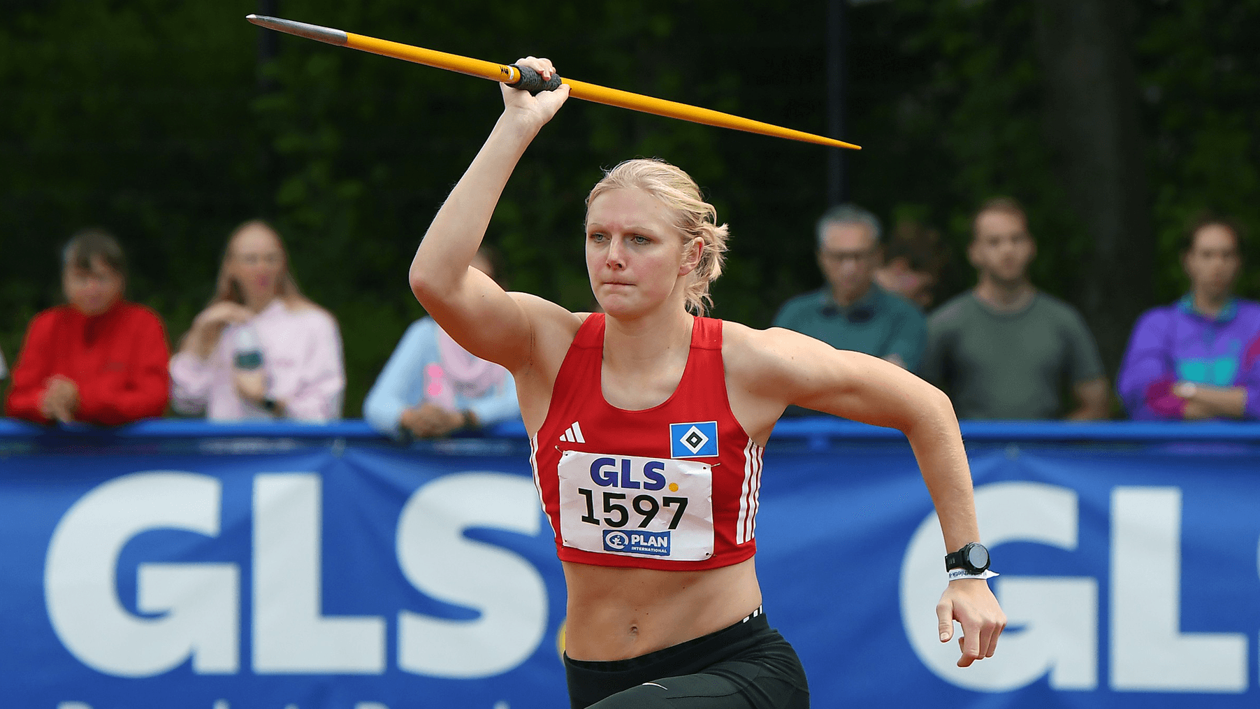 An athlete in a red tank top prepares to throw a javelin during a competition, with spectators in the background.