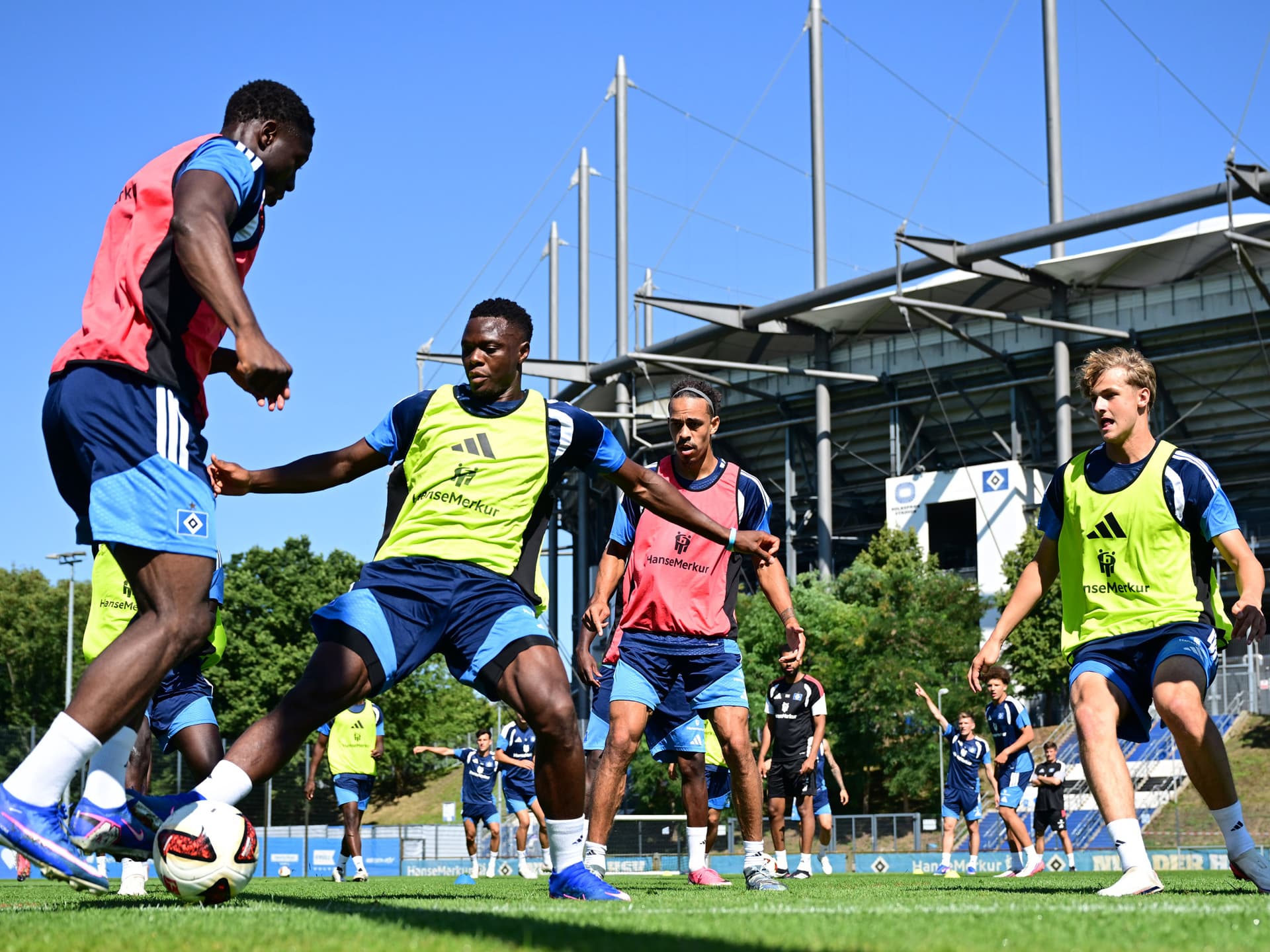 Kelvin Ojo am Ball im Training, ihm gegenüber Patson Daka und Louis Lemke, die den Ball attackieren, sowie im Hintergrund Yussuf Poulsen als Anspielstation. Insgesamt sommerliches Flair mit Volkspark im Hintergrund und kurzen Trainingsoutfits.