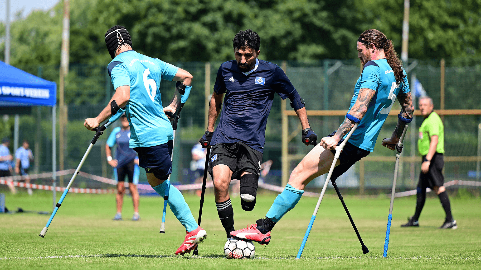 Three athletes playing amputee football fiercely contest the ball on a grassy field, all supported by crutches.