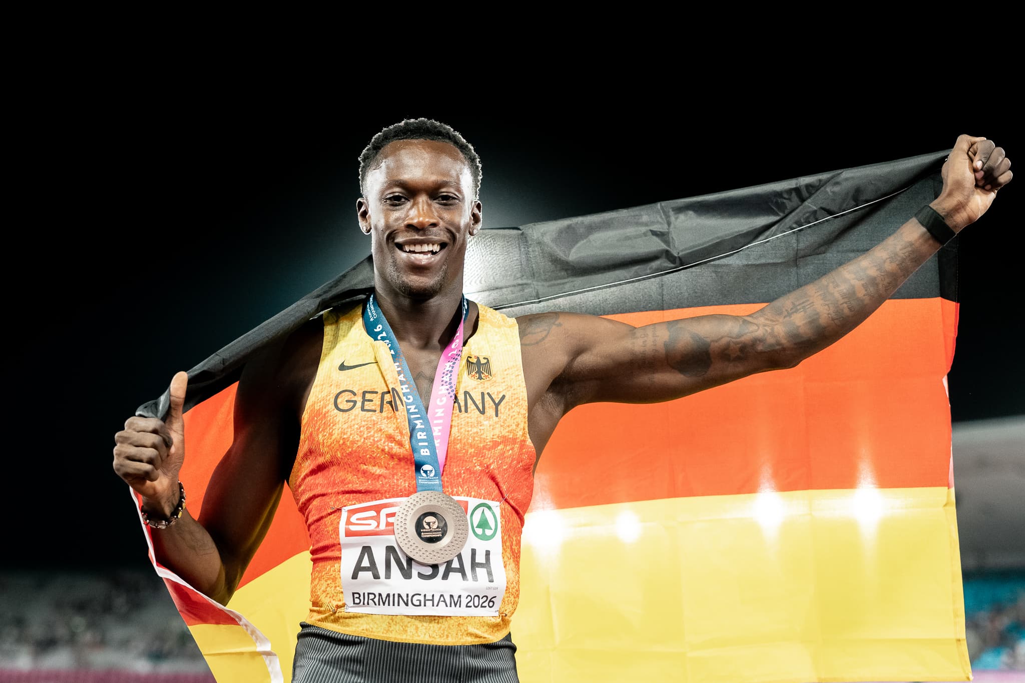 Athlete smiling, holding German flag, wearing a medal with "Birmingham 2026" and "GERMANY" on his shirt.