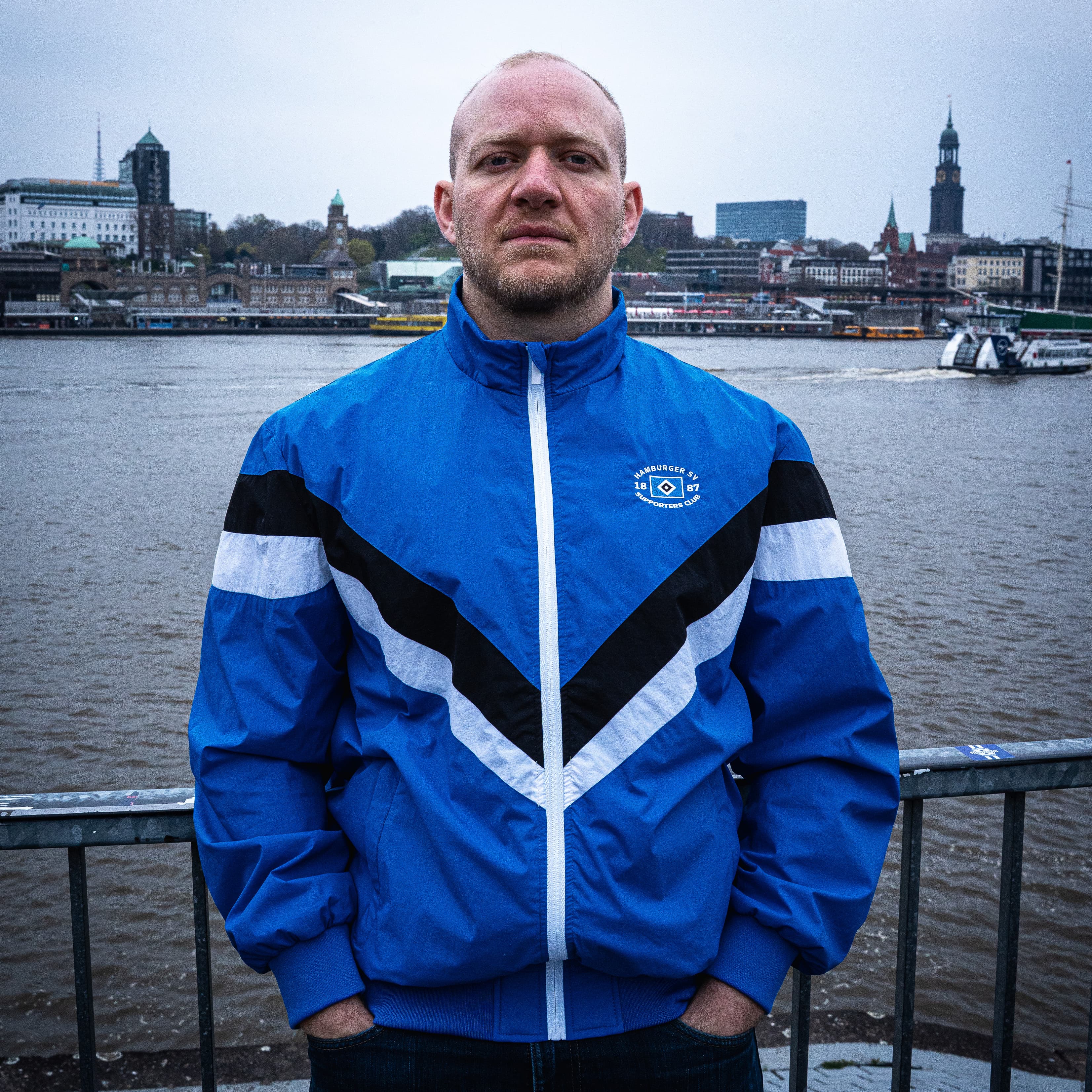 Man in a blue jacket stands by a river with an urban skyline in the background, featuring a prominent clock tower.