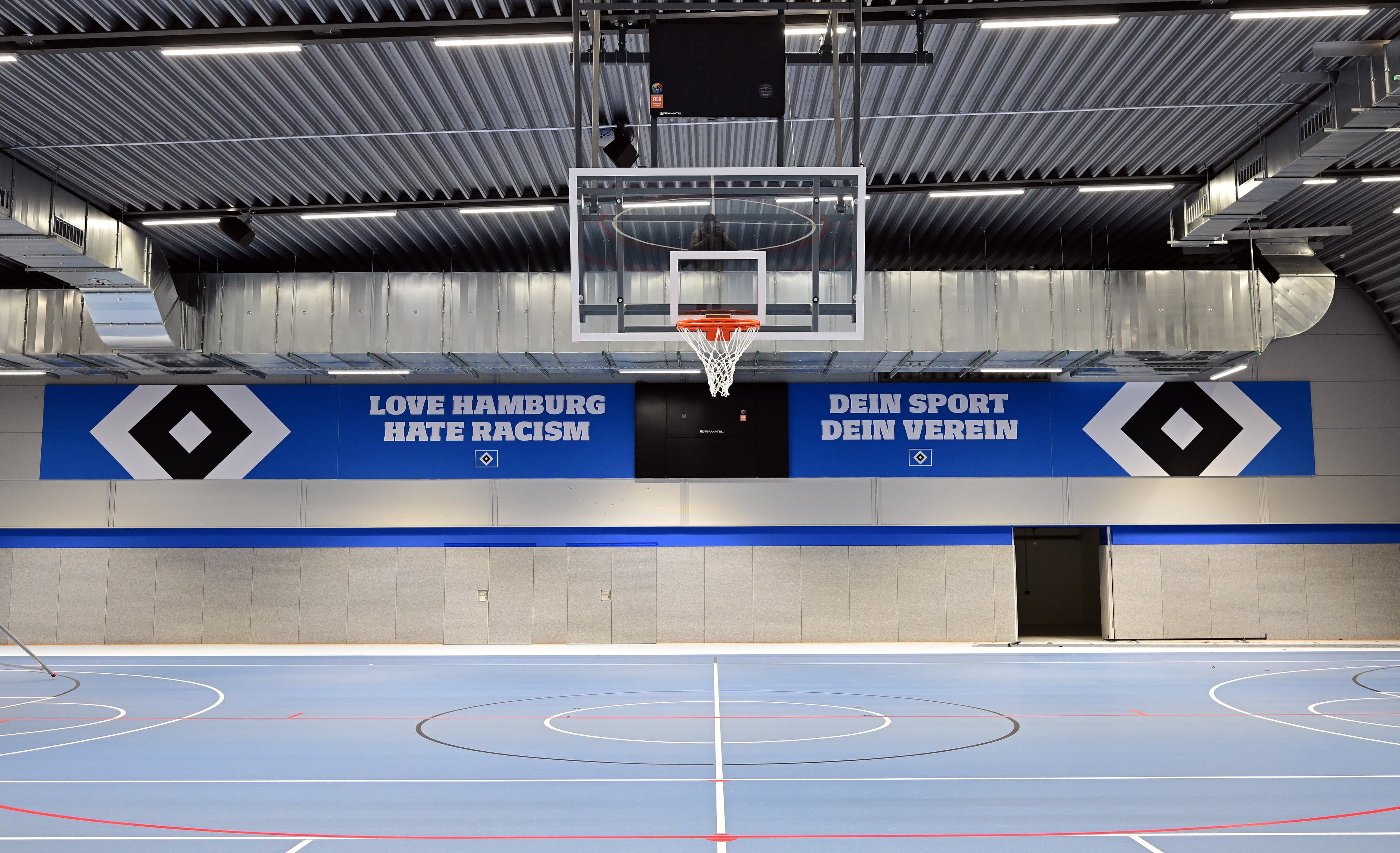 Indoor sports hall with a basketball hoop, blue and gray walls, and banners reading "Love Hamburg, Hate Racism" and "Dein Sport, Dein Verein."