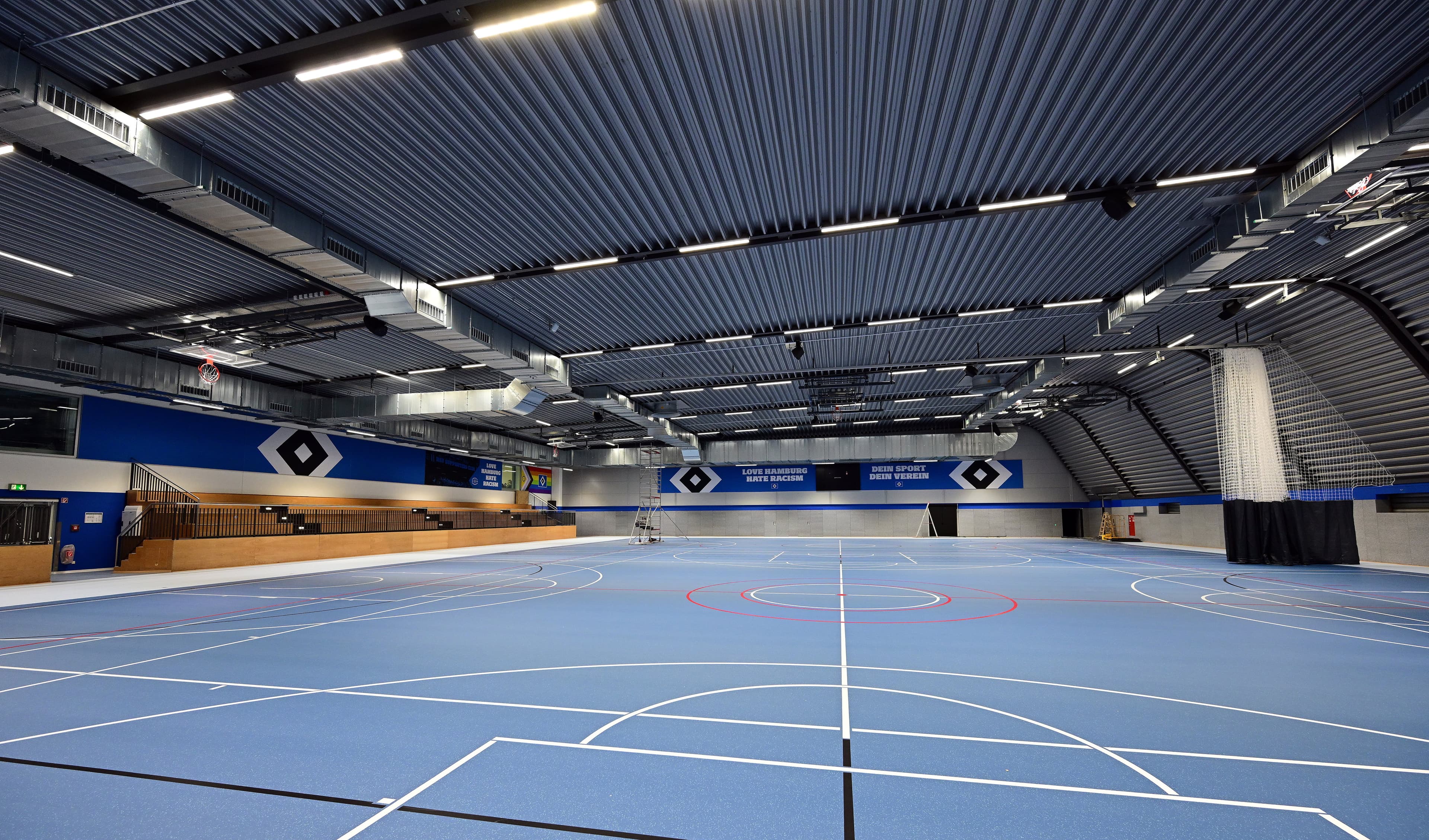 Indoor sports hall with blue flooring, marked with white and red lines for various sports; metal ceiling with ductwork and seating area on the side.