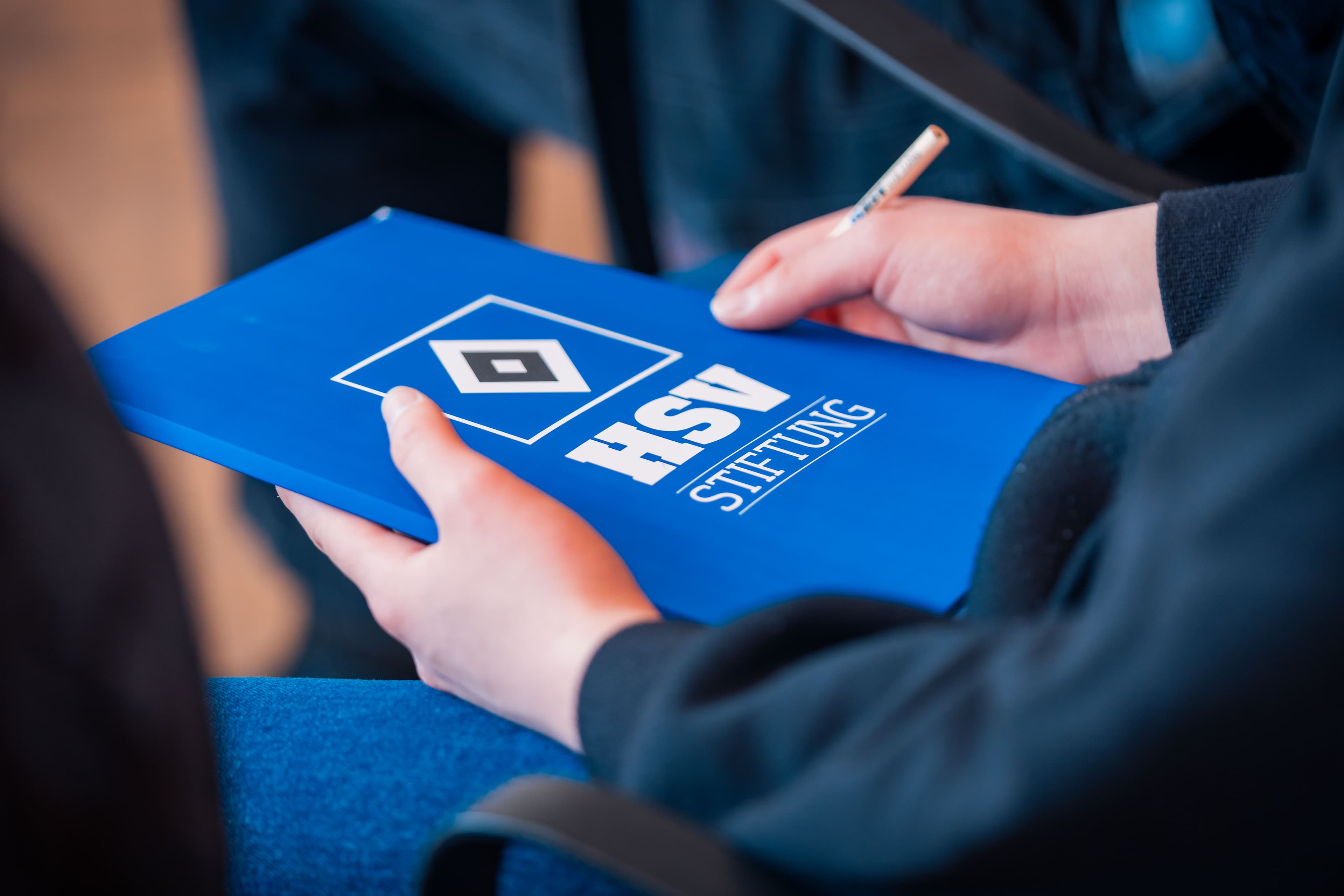 Ein Kind hat eine blaue HSV-Stiftung-Mappe und einen Stift in den Händen.