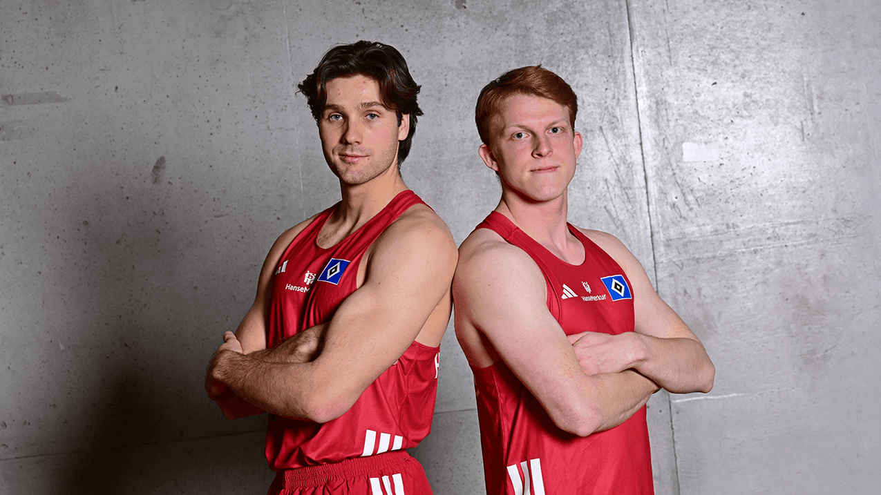 Two young men in red sports uniforms with arms crossed, standing back-to-back against a concrete wall.