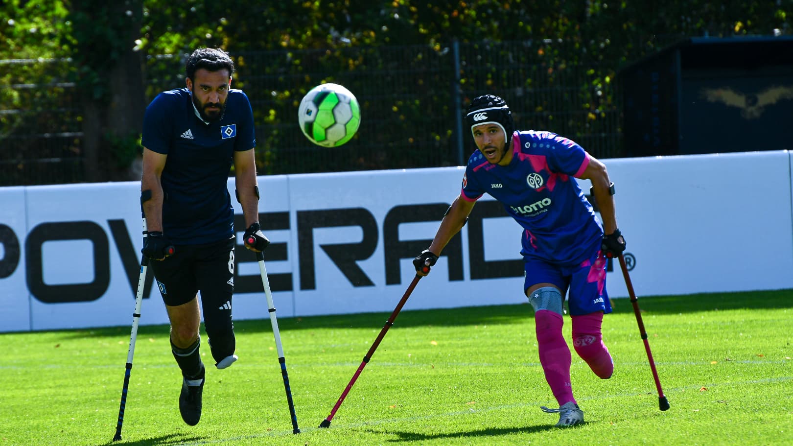 Two athletes on crutches playing soccer, one in a blue jersey and the other in a pink jersey, focus on a soccer ball mid-air.