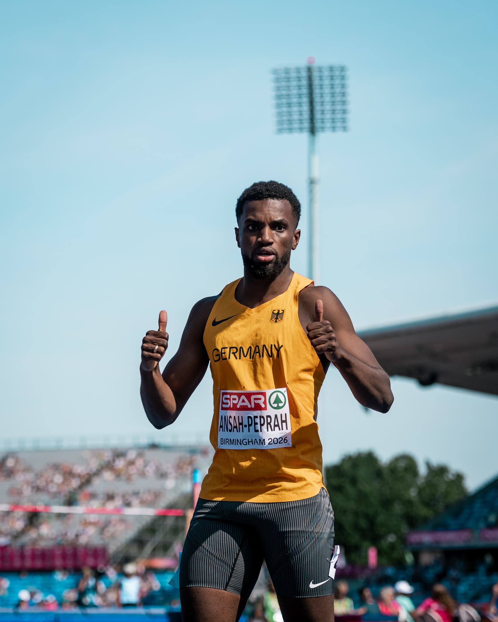 Athlete in a German track uniform gives thumbs up during an outdoor event, with a stadium and clear sky in the background.