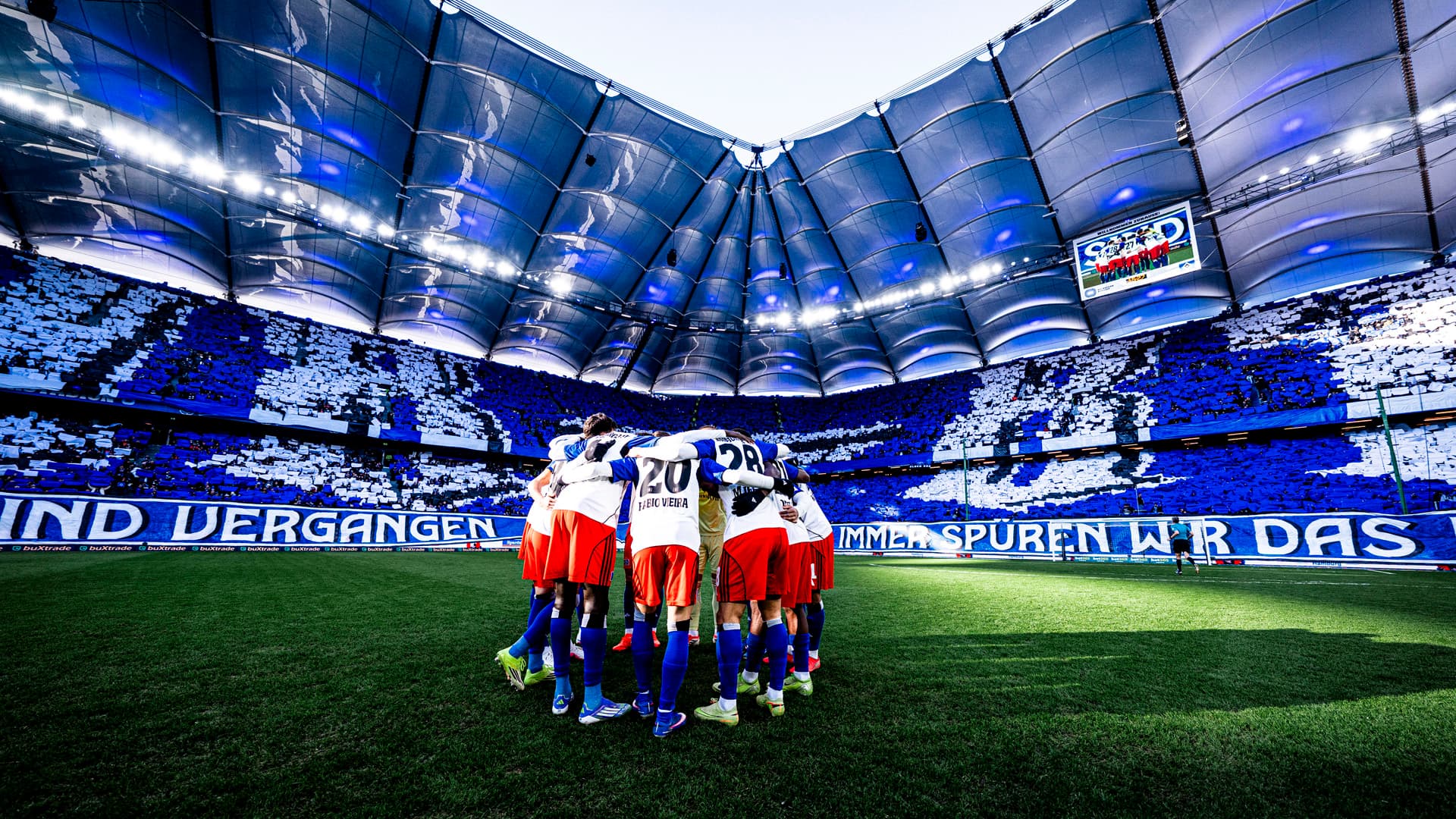 HSV team huddles on a lush green in the Volksparkstadium, surrounded by a full choreo of blue and white stands, under a bright, domed roof.