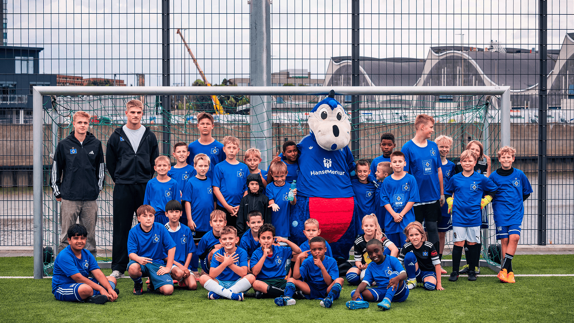 Eine Gruppe von Kindern posiert mit Dino Hermann vor einem Fußballtor.