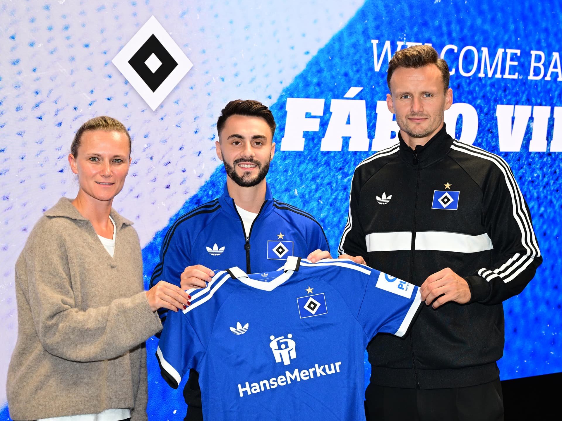 Kathleen Krüger, Fabio Vieira und Claus Costa halten ein HSV-Trikot hoch. Im Hintergrund steht auf der Rückwand "Welcome back, Fabio Vieira".