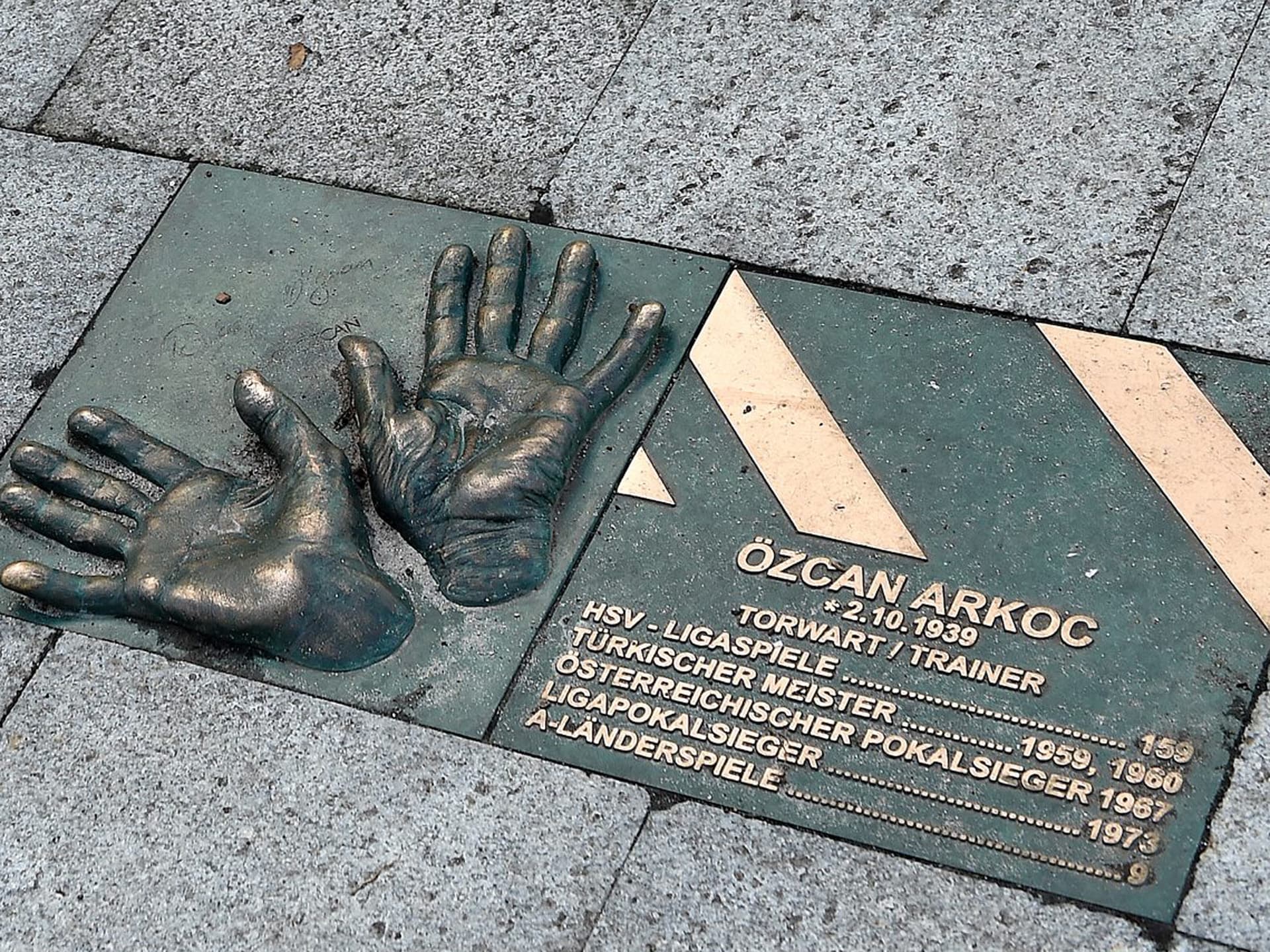 Bronzene Handabdrücke und eine Gedenktafel zu Ehren von Özcan Arkoc mit Informationen über seine Erfolge als Fußballtorwart und -trainer, eingelassen in einen Pflasterstein.