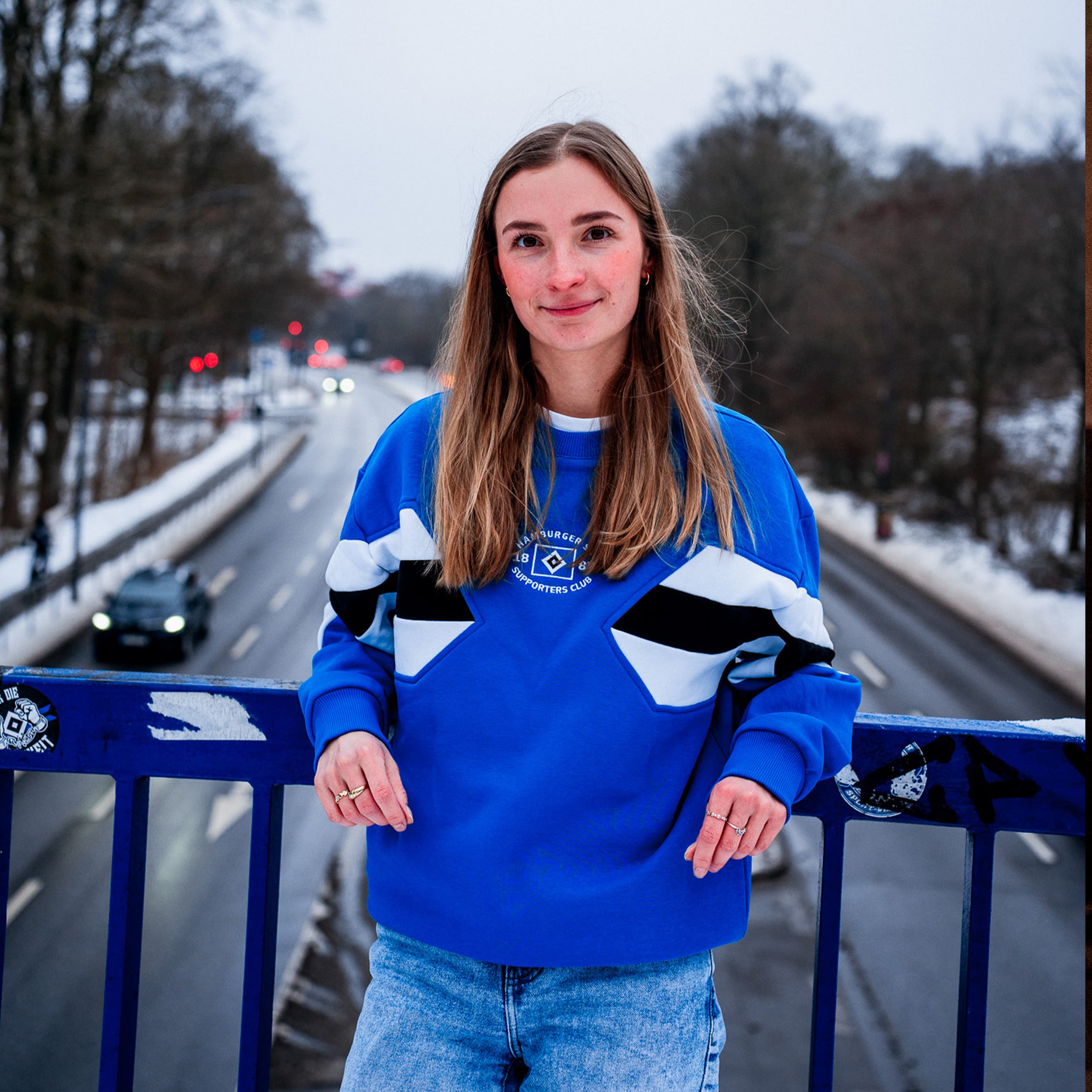 A woman in a blue and white sweater stands on a bridge with snowy trees and a road in the background.