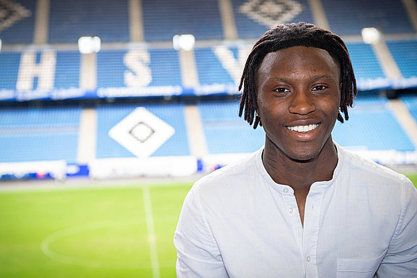 Mit einem Profispiel im Volksparkstadion würde für Opoku, der seit 2011 für den Verein spielt, ein Traum in Erfüllung gehen. Opoku lächelt und schaut in die Kamera. Im Hintergurnd ist das Innere des Volksparkstadions zu sehen.