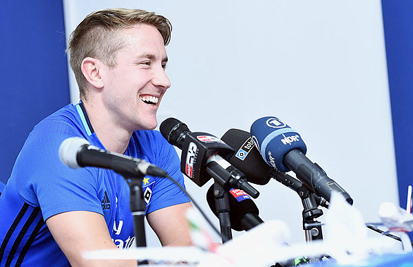 Lewis Holtby sitzt auf dem Podium vor zahlreichen Mikrofonen.