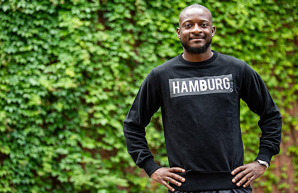 David Kinsombi steht im HSV-Pullover vor einer Hecke. 